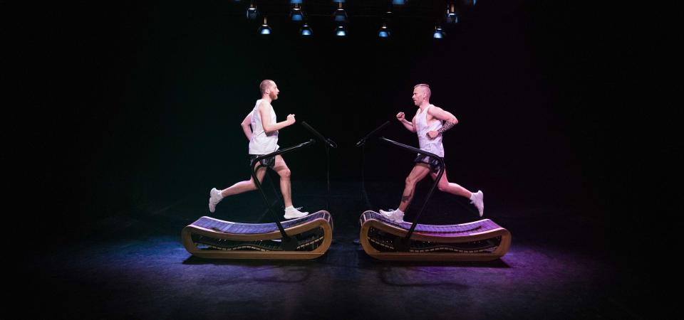 Man sieht zwei gegenüberstehende Laufbänder auf denen jeweils eine Person läuft, die beiden Personen laufen aufeinander zu. Der Hintergrund ist schwarz die zwei Personen sind lila und blau angeleuchtet